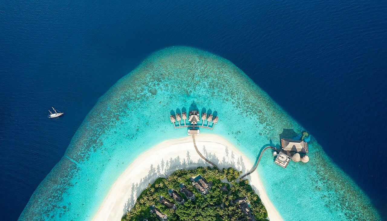 An aerial view of Anantara Kihavah Maldives Villas reveals a vibrant turquoise lagoon encircling the island, framed by soft white sands and dense tropical greenery. Private Beach Pool Villas are nestl