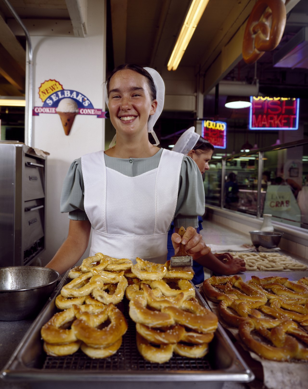 Title: A young Amish woman from Lancaster County serves fresh-cooked soft pretzels, a time-honored Philadelphia delicacy at the Reading Terminal Market in Philadelphia, Pennsylvania
Physical descripti