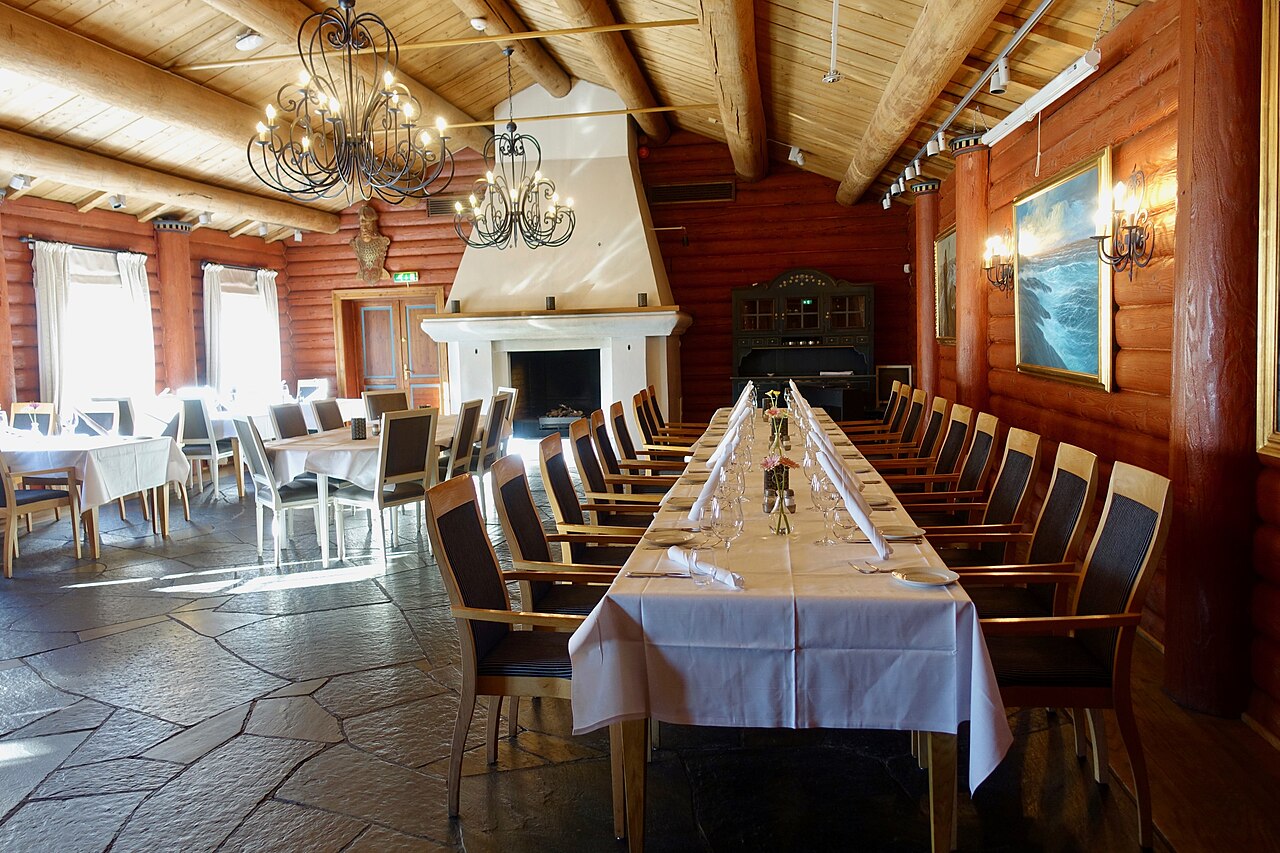 Interior of Leangkollen Hotel in Asker, Norway. Dining hall with log walls, maritime paintings, dining tables, wrought iron chandeliers, sunlight, fire place, etc. 
The conference hotel was originally