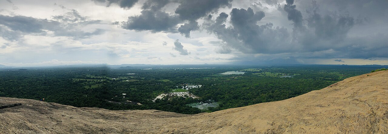 Panoramic view of Sigiriya rock fortress seen from neighboring Pidurangala Rock, surrounded by jungle and paddy fields.
