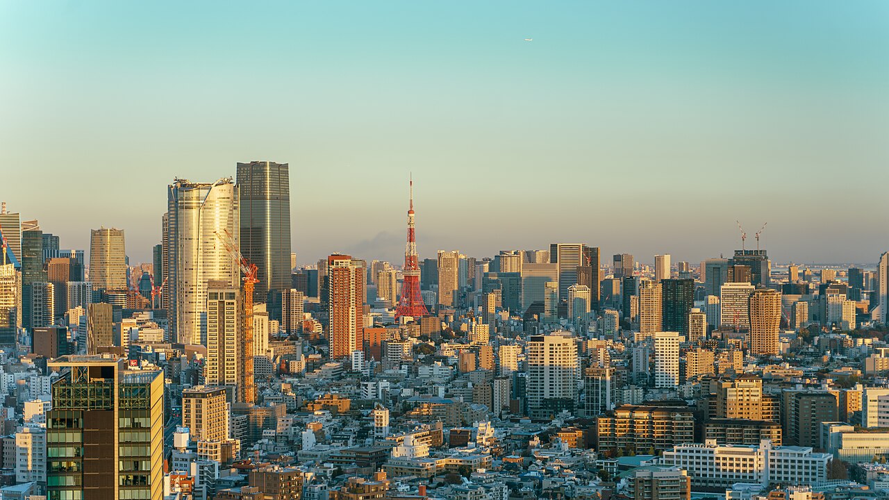 A wide-angle landscape photograph of the Tokyo skyline captured during the "golden hour." The image features a dense concentration of skyscrapers in the Minato ward. The iconic Tokyo Tower, a communic